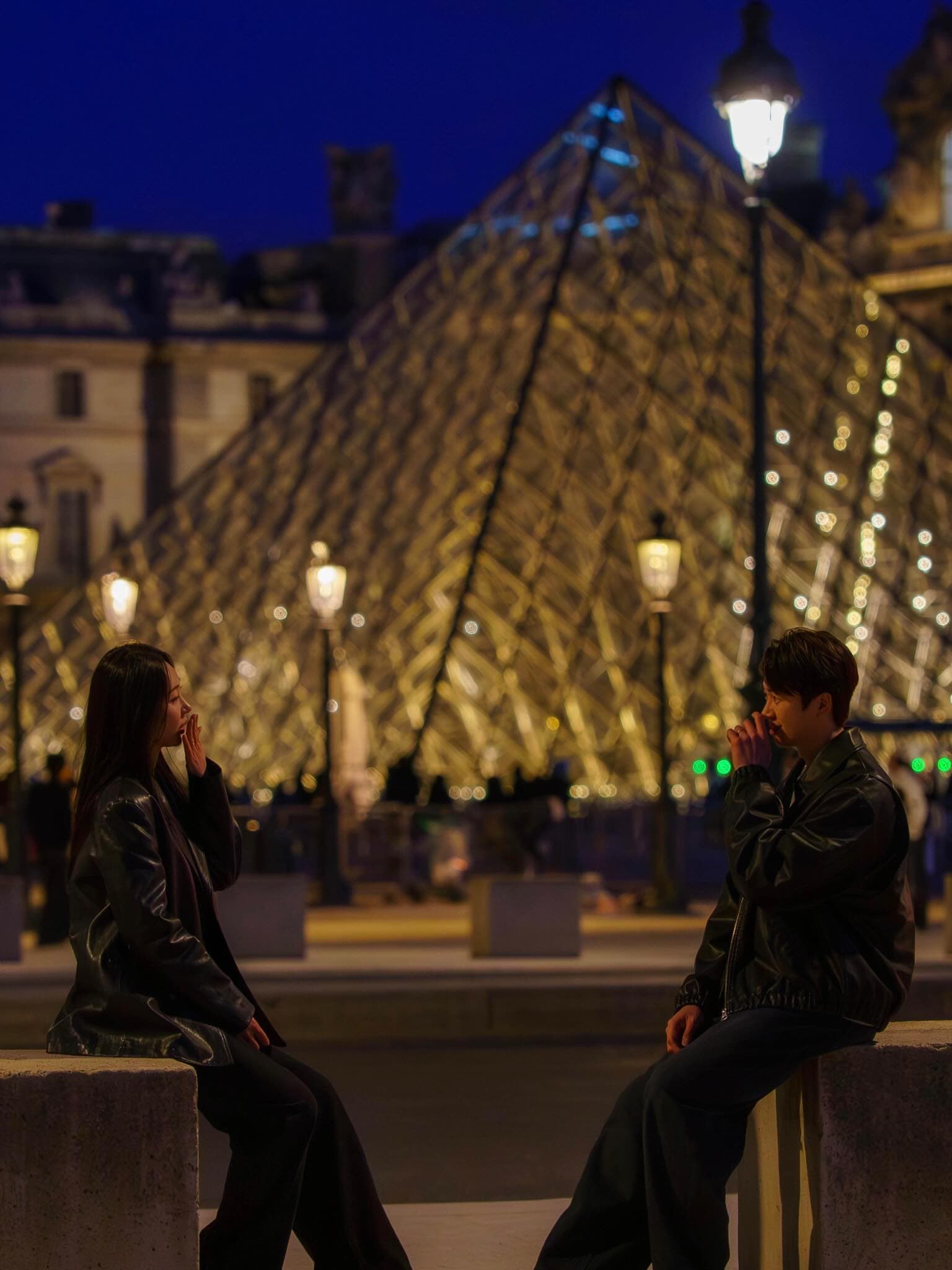 밤의 루브르 유리 피라미드 앞에 앉은 커플의 실루엣